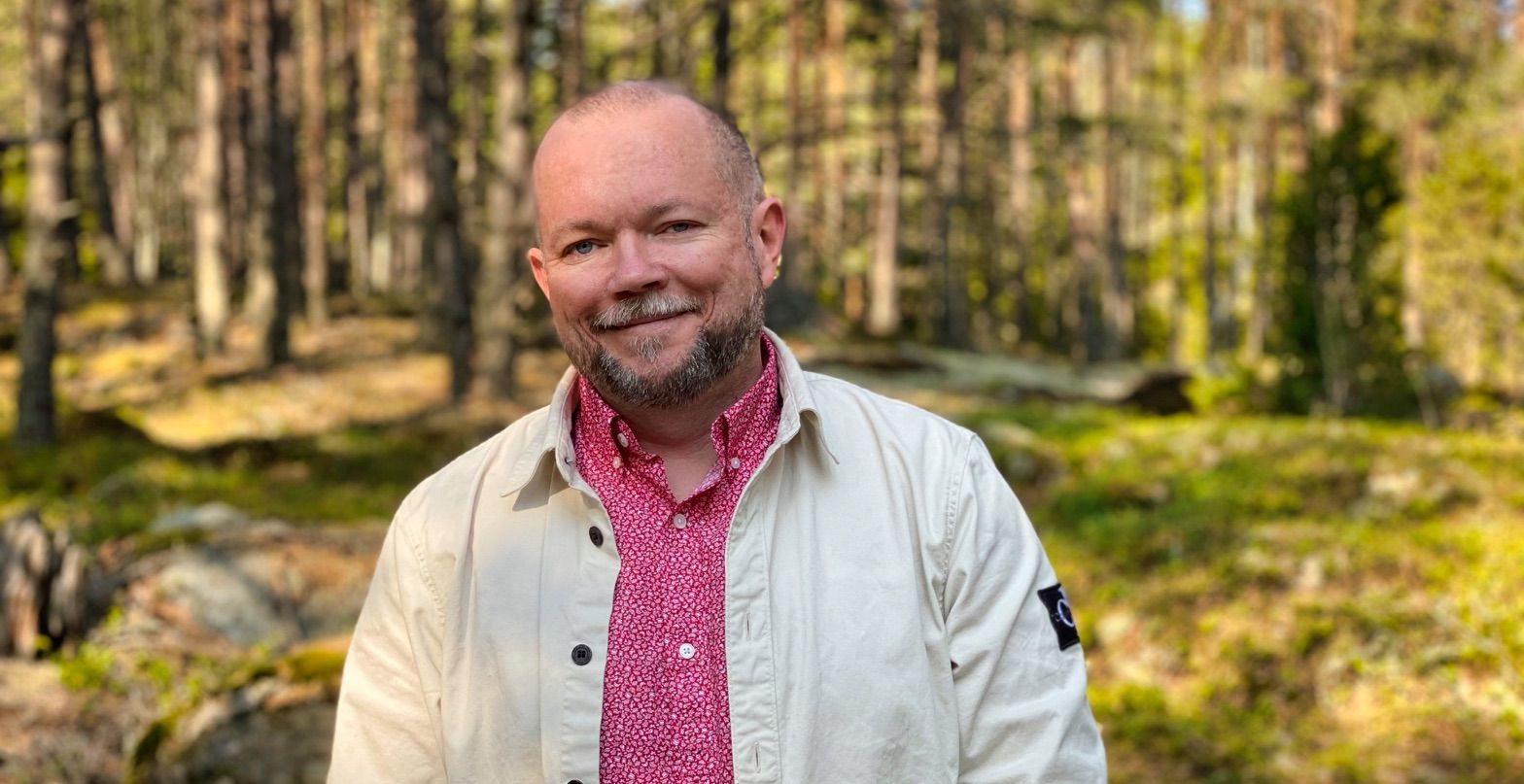 Per Axbom med röd skjorta, beige overshirt och leende, med en skogsmiljö i bakgrunden.