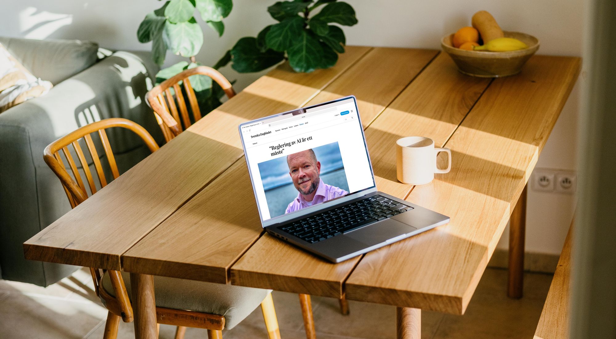 Köksbord i trä. På bordet står en bärbar dator där Pers debattartikel syns på skärmen. En vit kaffemugg står bredvid datorn.