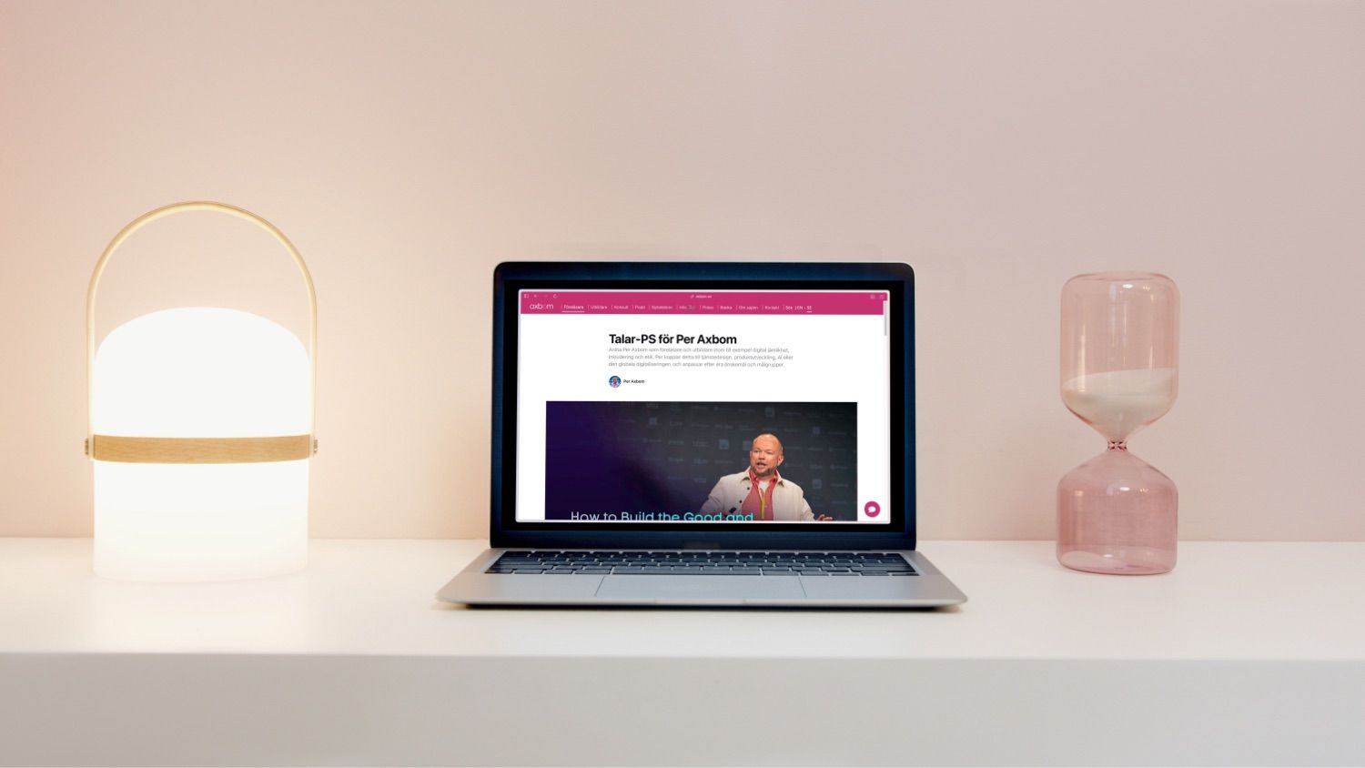 Webbplatsen axbom.se syns på en laptop som står mot en ljus avskalad vägg. En lampa och ett timglas står på vardera sida.