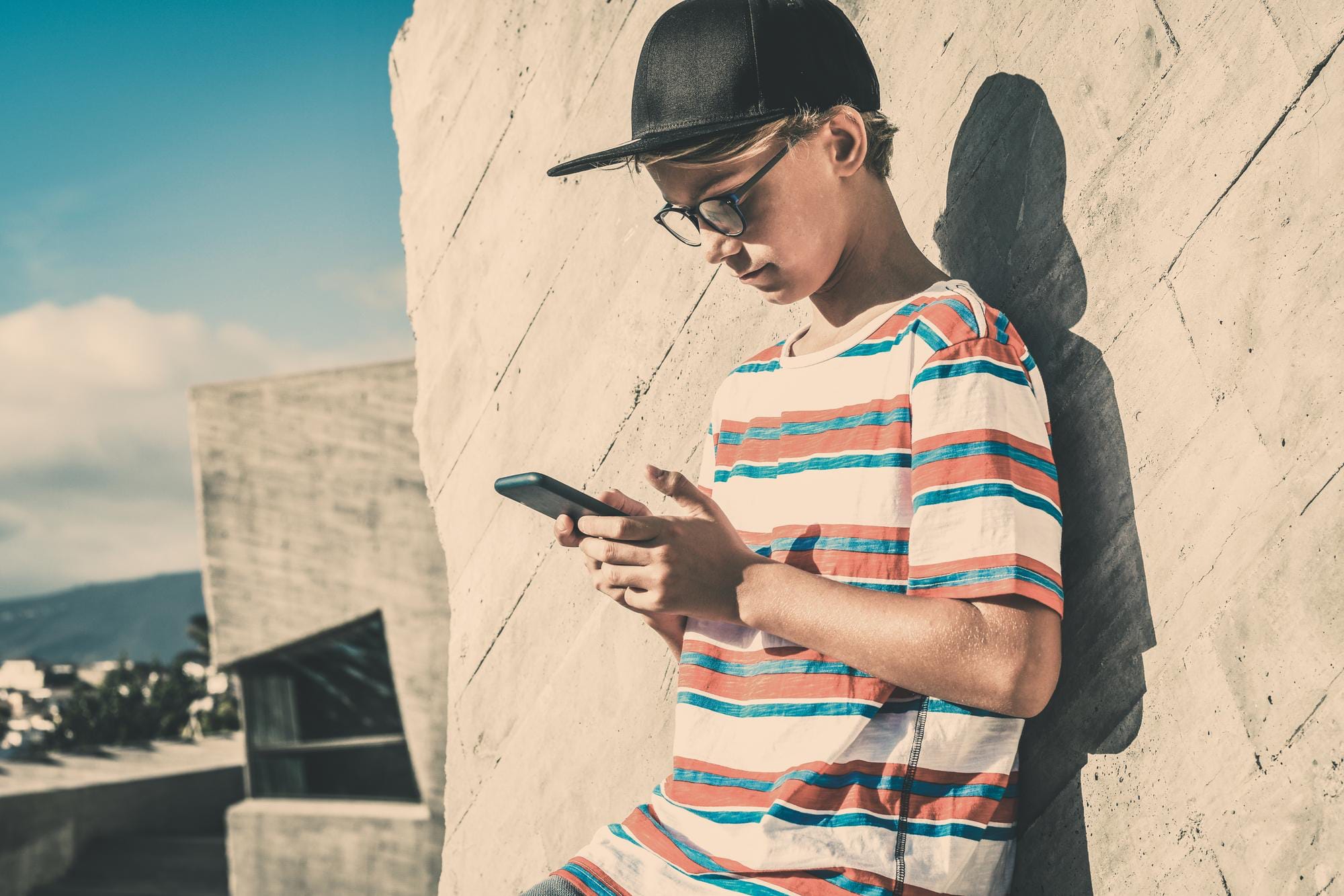 Ungdom i keps och glasögon som lutar sig mot en stenvägg och skriver på en mobil.
